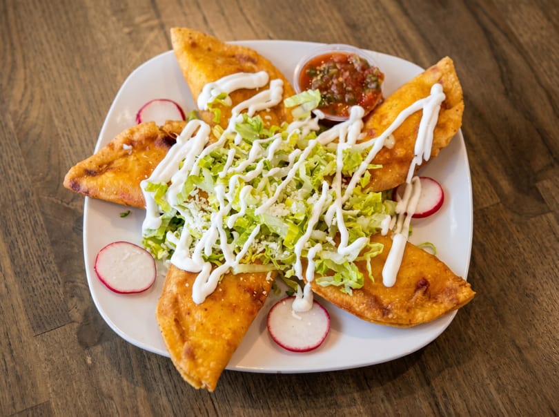 Four golden-fried quesadillas topped with shredded lettuce, sour cream, and radishes, served with salsa.