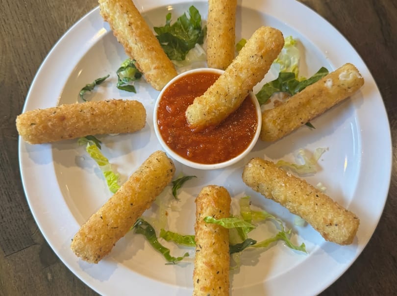 Golden-brown mozzarella sticks arranged around a bowl of red marinara sauce on a white plate.