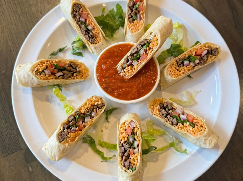 Seven mini burritos filled with meat, rice, and vegetables, arranged around a bowl of salsa.